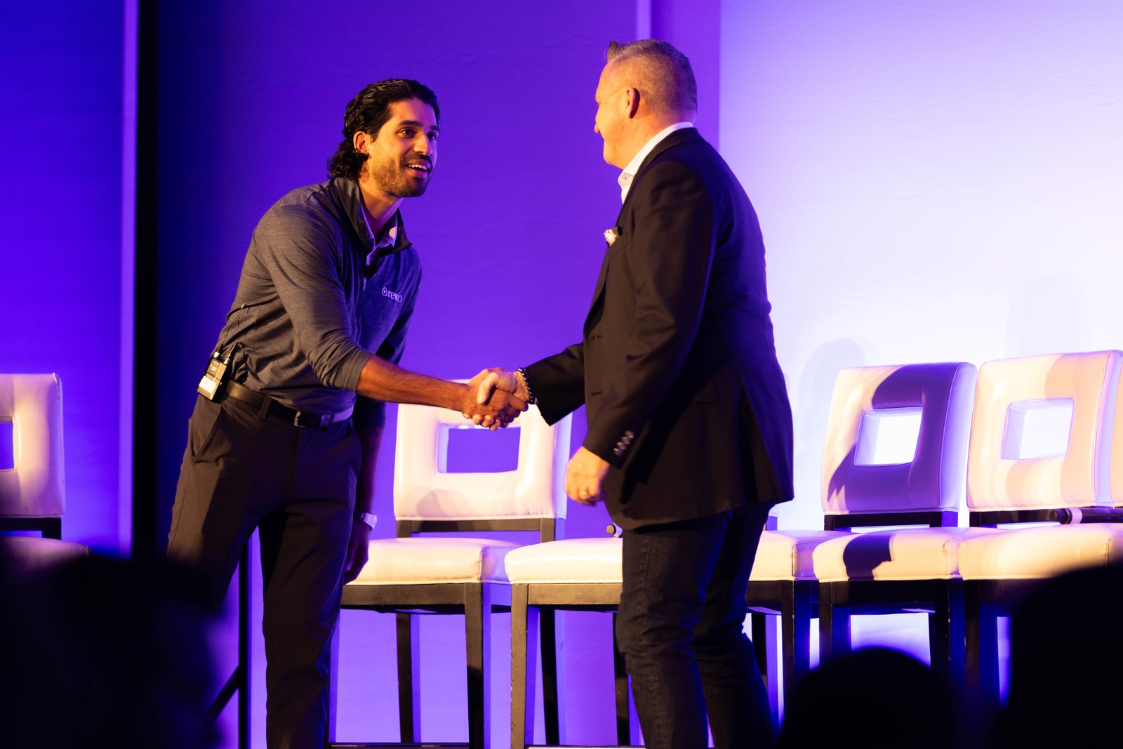 A Rev.io team member shakes hands with a guest speaker on the main stage under purple lighting
