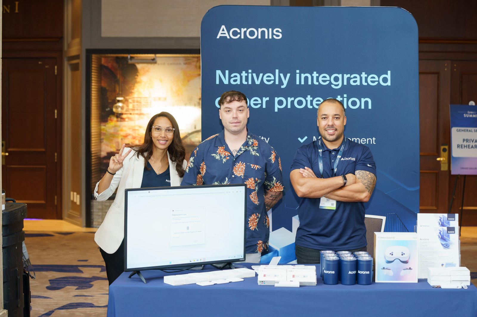 Three Acronis team members at their booth in the Summit expo hall with a banner reading Natively Integrated Cyber Protection