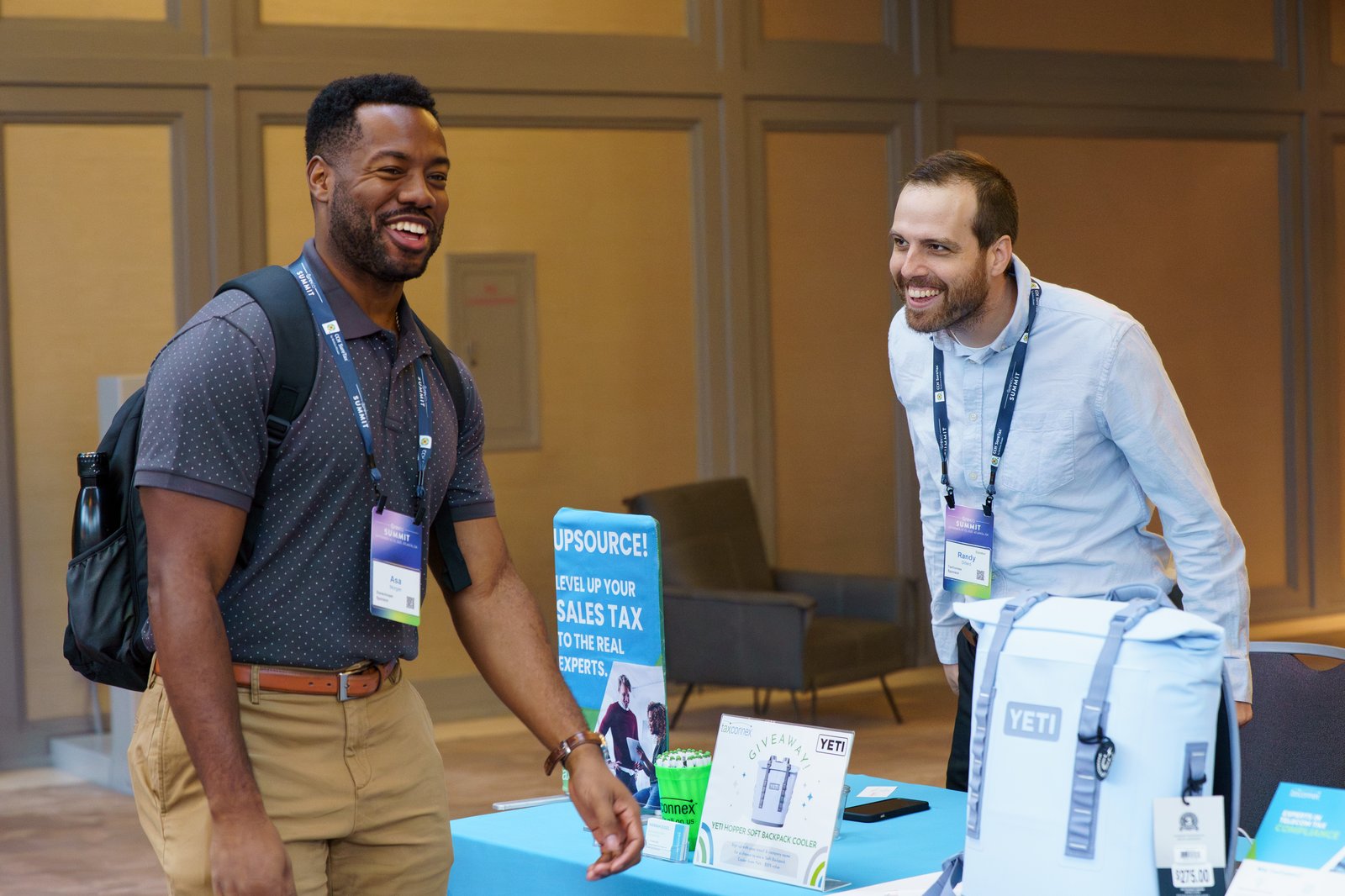 Two Summit attendees laughing at a TaxConnex sponsor booth featuring a YETI cooler giveaway