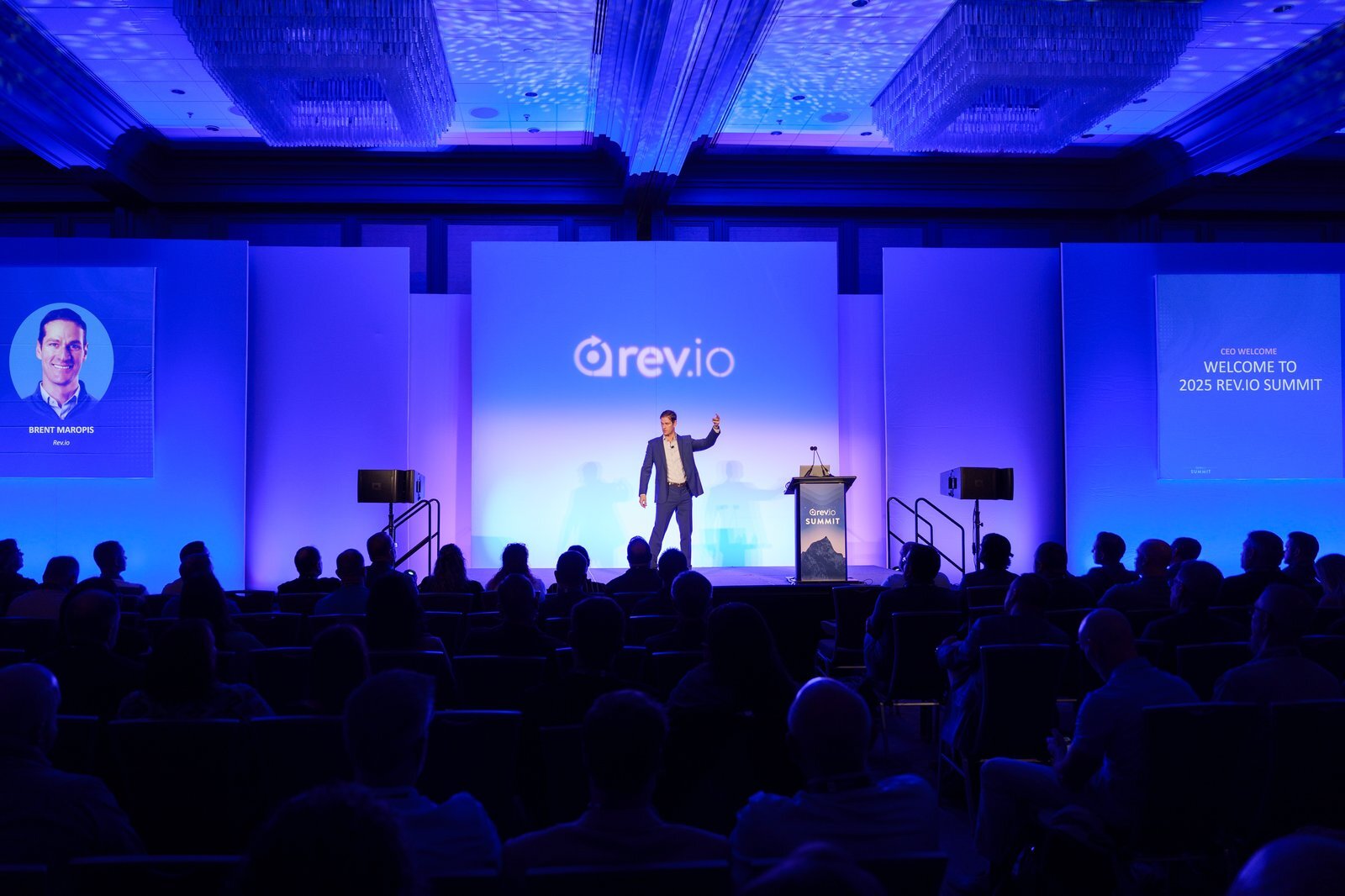 Rev.io CEO Brent Maropis on the main stage opening Summit 2025 with a welcome slide projected behind him