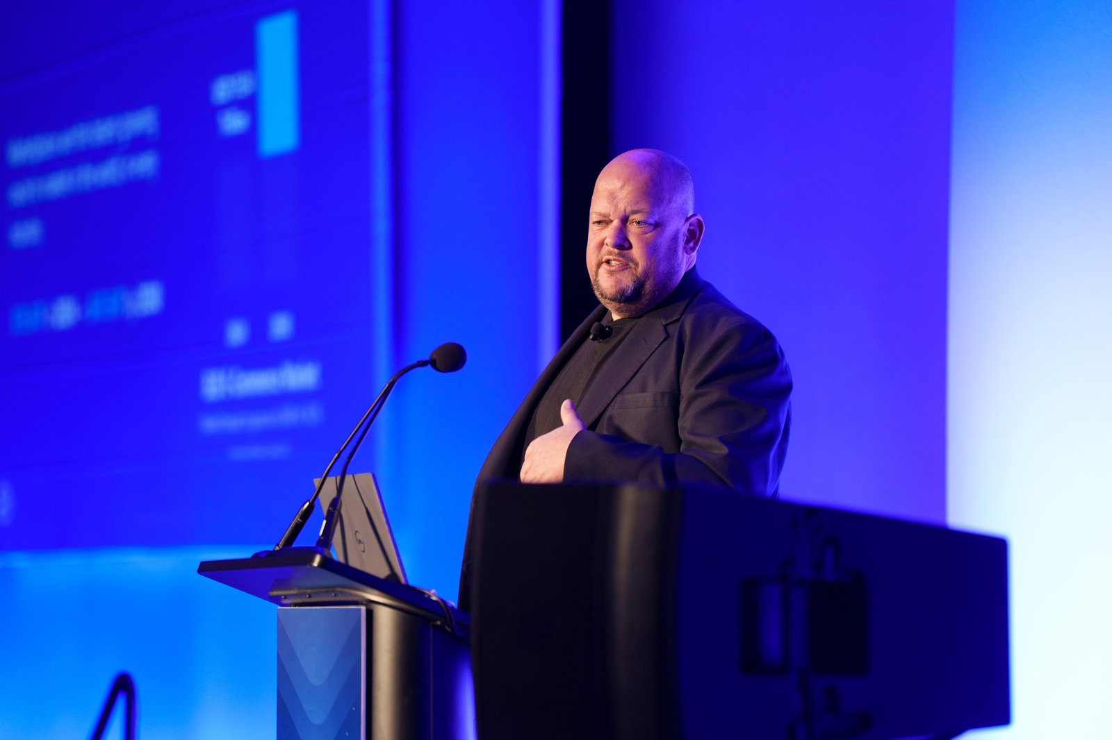 A keynote speaker at the podium in a black blazer presenting under blue stage lighting