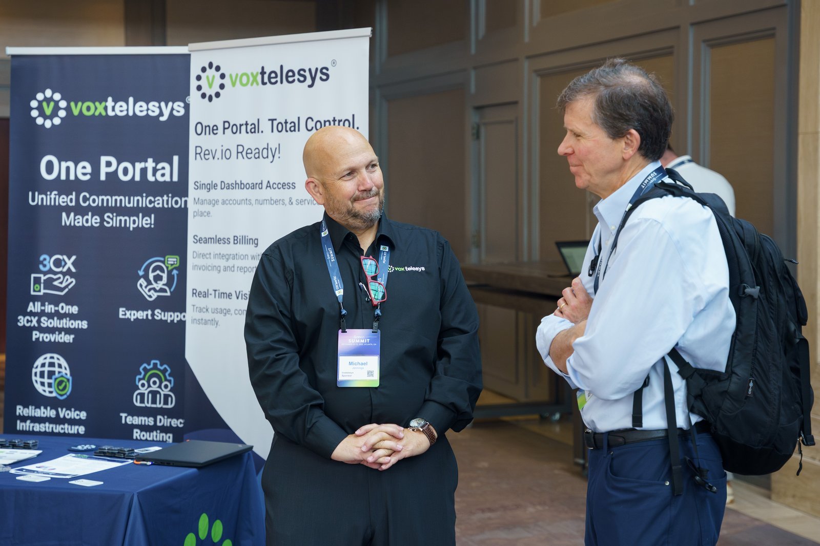 A Voxtelesys representative talking with an attendee in front of two One Portal Total Control banners in the expo hall