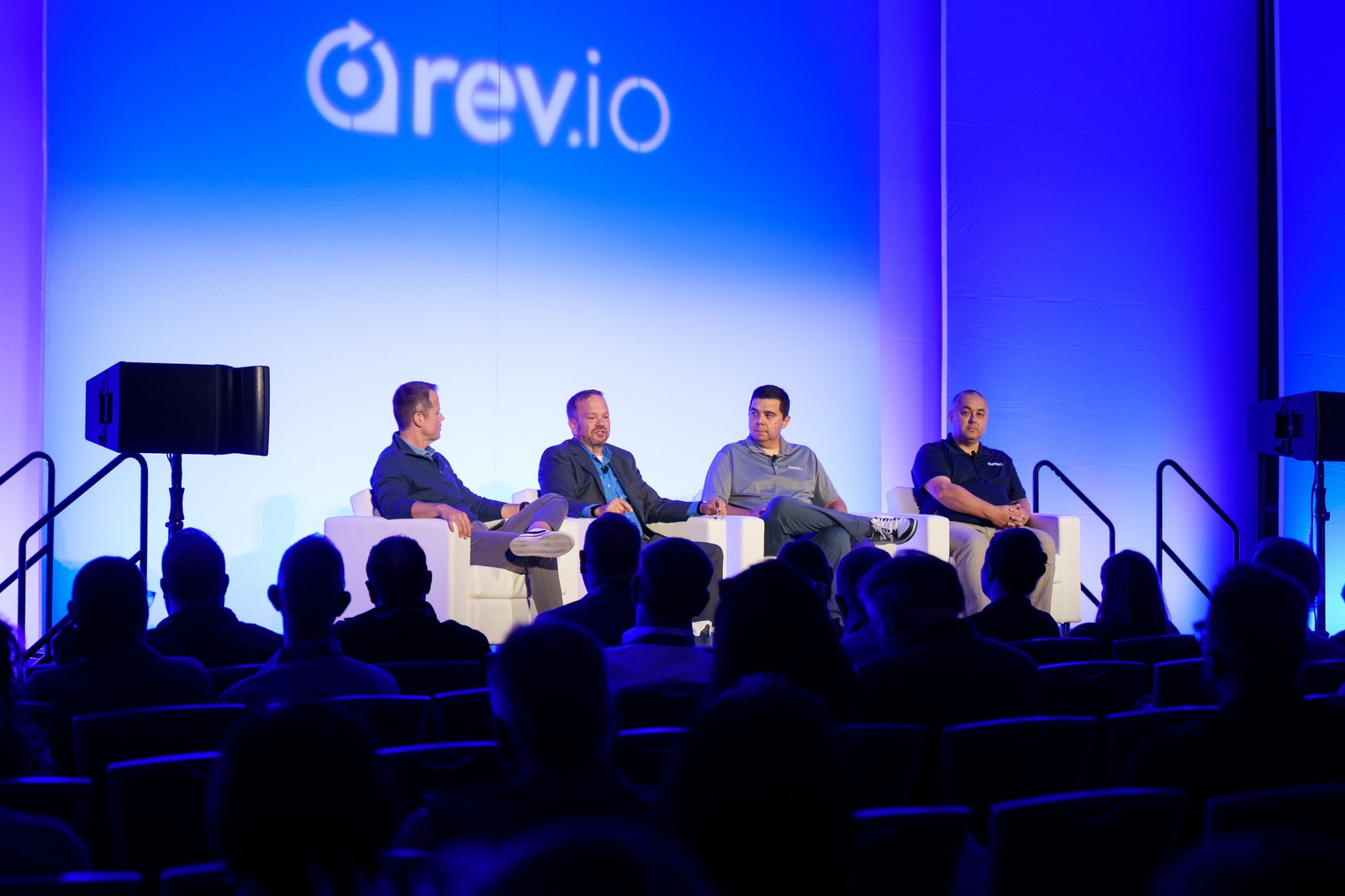 Four panelists seated in white chairs on the Grand Ballroom stage during a Summit 2025 session with the Rev.io logo projected behind them
