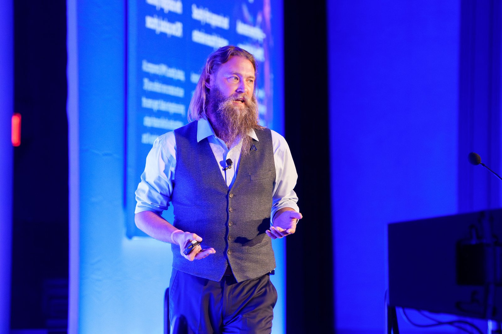 A speaker in a gray vest and blue shirt presenting a deep dive session under blue stage lighting